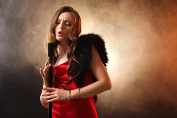 Beautiful young woman in stylish dress with microphone singing on dark background in color lights and smoke