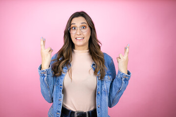 Young beautiful woman wearing denim jacket over isolated pink background gesturing finger crossed smiling with hope and looking side