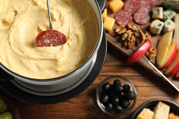 Fork with piece of sausage, melted cheese in fondue pot and other products on wooden table, flat lay