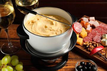 Fork with piece of sausage, melted cheese in fondue pot and other products on wooden table, closeup