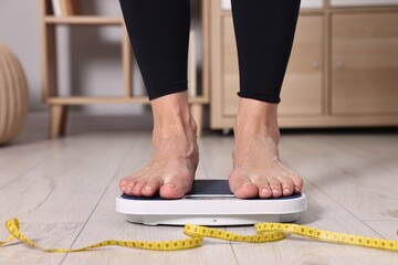 Woman standing on floor scale and measuring tape at home, closeup. Weight control