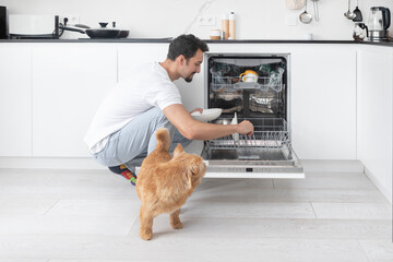 Young man loading dirty dishes inside dishwasher machine and his cute dog trying to play with him	