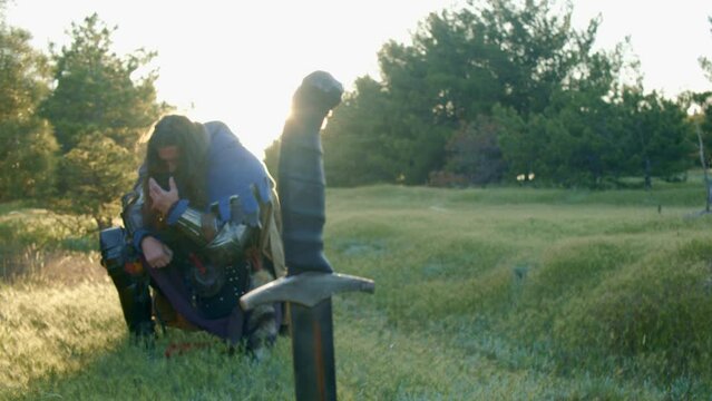 A man with a beard in a medieval knight costume stands kneeling in front of a sword stuck in the ground in the bright rays of the sunset. Pose of grief and thirst for revenge. Close-up