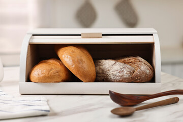 Wooden bread basket with freshly baked loaves and spoons on white marble table in kitchen