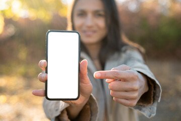 Woman using smartphone in park, pointing finger at blank white mobile phone screen