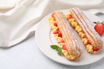 Delicious eclairs filled with cream, strawberries and mint on white tiled table, closeup