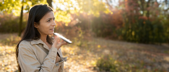 Hispanic woman using voice assistant on mobile phone, talking to smartphone walking in autumn forest