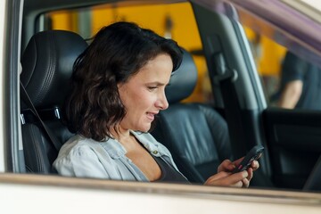 Female customer in blue denim shirt sitting in car, smiling and looking at smartphone. Background includes garage setting. Represents communication or transaction related to car repair services. - Powered by Adobe