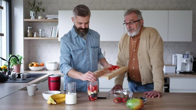 Senior father and mature son making fruit smoothie as snack, having fun in kitchen. Handsome man pouring milk into blender with fruits.