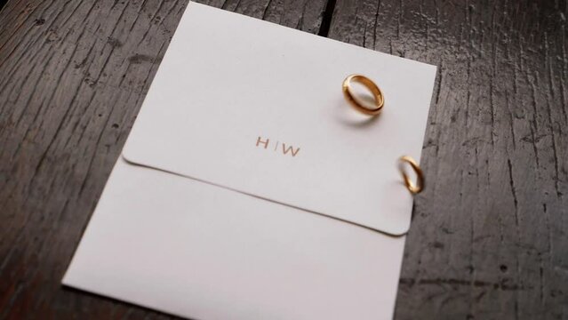 gold wedding rings spinning on invitation with initials H and W written in gold on wooden boards. Jewelry for a married couple. 60fps