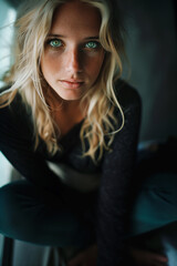 Fototapeta premium A young woman with blonde wavy hair and green eyes, sitting casually, her face adorned with freckles. She gazes intensely, bathed in soft, natural light.