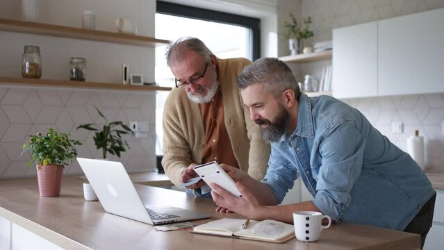 Mature son helping father to manage his finance, teaching how to work with internet baking and shopping online. Handsome son supporting dad, technology and digital literacy.