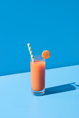 Jugo de zanahoria en un vaso transparente con pajita adornado con rodaja de zanahoria sobre fondo azul