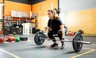 A middle-aged adult woman is about to lift a barbell with metal discs to do a strength...