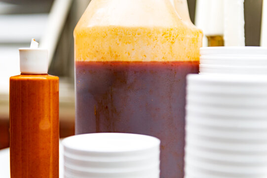 Nondescript containers of barbecue sauces on a table next to stacks of white cups