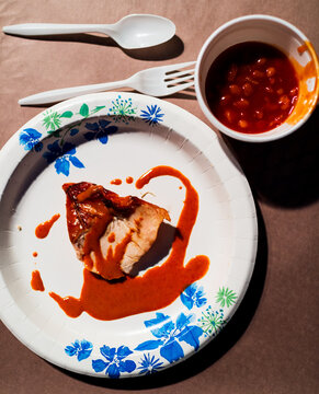 A large slice of cooked pork doused in reddish barbecue sauce on a white paper plate with floral designs. Next to it are white plastic cutlery and a styrofoam cup of baked beans