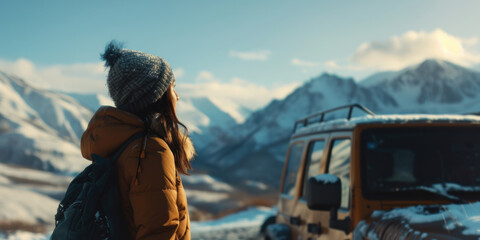 A woman wearing a hat and a backpack stands in front of a yellow jeep. The scene is set in a snowy mountain range, and the woman is looking at the jeep