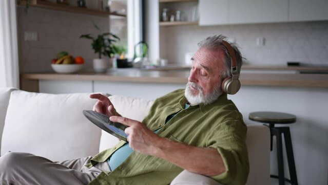 Elderly man listening to vinyl with headphones, sitting, relaxing on sofa. Pairing turntable with pair of wireless headphones at home.