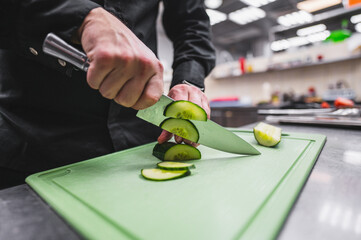 A person skillfully slices a fresh cucumber on a green cutting board using a chef’s knife