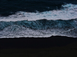 Aerial photo of a sea water surface. Blue water with foam on waves. Beach with black volcanic sand - view from above. Stylized background abstract texture photo. Tenerife, Puerto de la Cruz