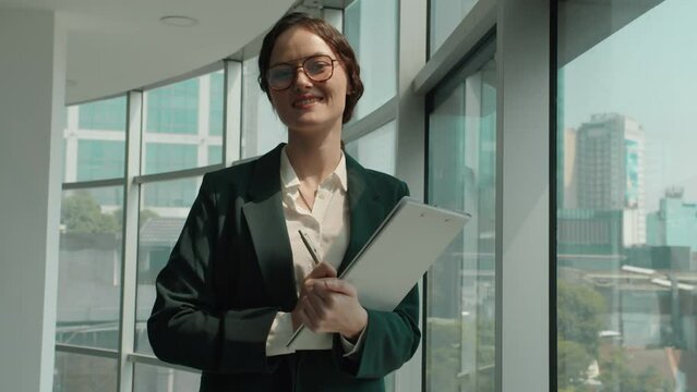 Medium portrait of young Caucasian businesswoman filling form in folder and looking at camera in modern coworking space