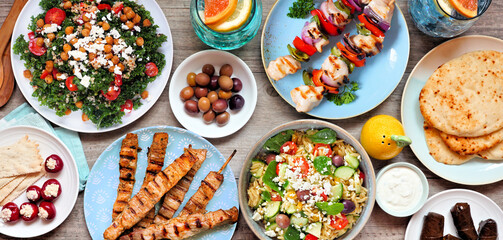Summer Mediterranean theme food table scene. Top view on a wood banner background. Grilled skewers, salads, snacks, pita and tzatziki.