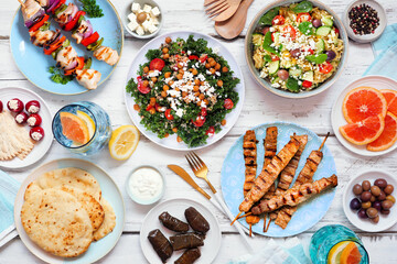 Summer Mediterranean theme food table scene. Above view on a white wood background. Grilled skewers, salads, snacks, pita and tzatziki.