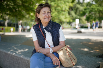 Elderly woman sitting on a park bench with a straw bag, smiling on a sunny day, enjoying the...