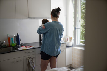 Woman in blue shirt holding baby in kitchen, multitasking between parenting and daily chores,...