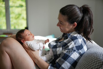 Mother cradling newborn baby in her lap, both sharing a loving gaze, indoor setting, warm and...