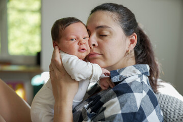 Mother holding newborn baby close to her face, both in a calm and intimate moment, indoor scene...