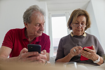 Elderly couple sitting together and using smartphones, focused on their screens, indoor scene with...