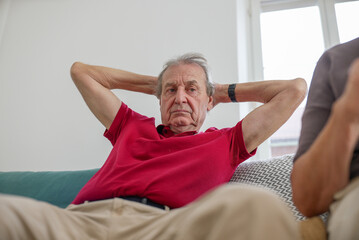 Elderly man relaxing on a couch with hands behind his head, indoor setting with natural light,...