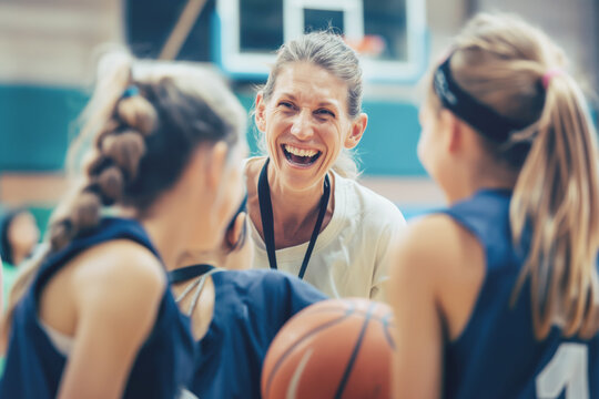 Laughing basketball coach with young female players during practice in gym. Joyful and energetic environment fostering teamwork and enthusiasm