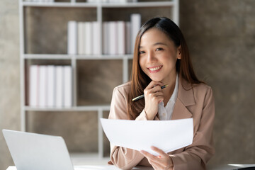 A happy businesswoman sits at her desk holding documents. She is smiling and appears satisfied, with a laptop and bookshelves in the background, creating a professional office setting.