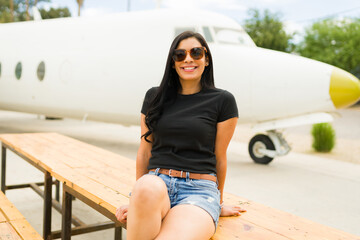 Fototapeta premium Happy latina woman relaxing on a bench, sporting sunglasses and a black t-shirt mockup, with a backdrop of a white airplane