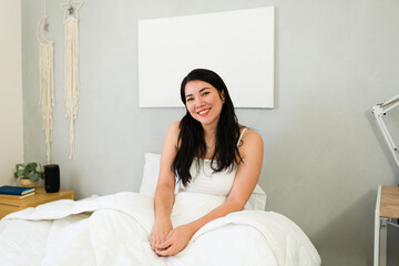 Happy woman with a charming smile lounges on her bed, radiating tranquility and joy in a peaceful bedroom