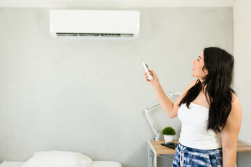 Happy woman adjusting mini split air conditioning unit with remote control on a hot summer day next...