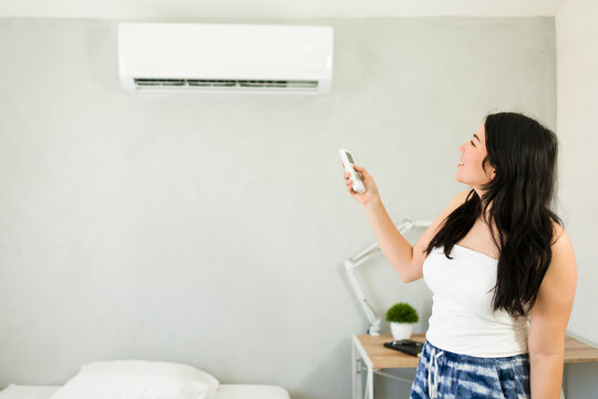 Happy woman adjusting mini split air conditioning unit with remote control on a hot summer day next to bed