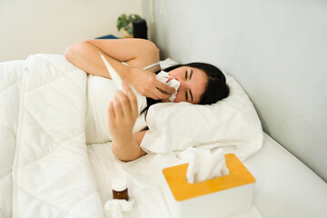 Sick young Caucasian woman in bed surrounded by tissues and medicine in a bedroom setting