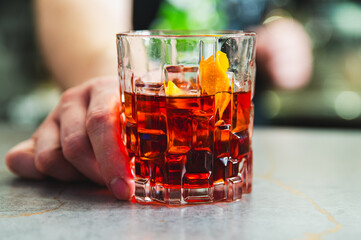 A close-up of a hand holding a glass of amber-colored liquid with ice cubes and an orange peel