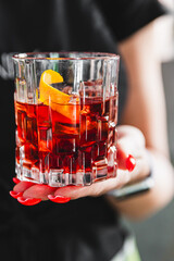 Close-up of a hand holding a glass of iced red beverage with an orange peel garnish