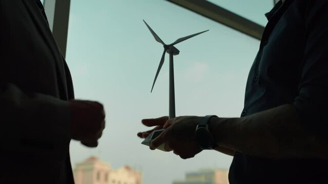 Low-key cropped shot of hands of two unrecognizable entrepreneurs discussing alternative energy implementation in their business, holding mockup of wind turbine