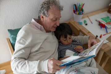 Elderly man reading a comic strip book with his young grandson in a cozy bedroom setting,...