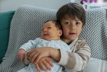 Young boy holding his infant sibling, both seated comfortably on a patterned cushion.