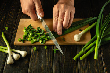 Chef hands using a knife to cut green garlic on a cutting board for preparing a vegetarian dish or...