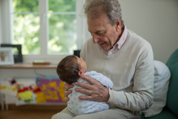 Grandfather and newborn sharing a heartfelt moment, with the elderly man gazing lovingly at the baby. The cozy indoor environment highlights their special bond and affection