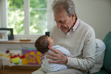 Old man with grey hair, holding an infant in his arms, looking down with a smile. This indoor scene...