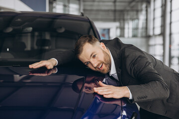 Positive young bearded caucasian man hugging his new car in dealership. Happy man finally gets...
