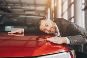 Positive young bearded caucasian man hugging his new car in dealership. Happy man finally gets...